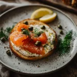 Freshly toasted bagel with cream cheese, smoked salmon, and capers on a wooden board.